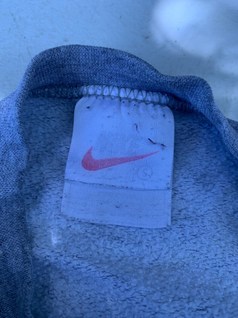 May include: Close-up of a gray sweatshirt collar with a white tag. The tag features a pink Nike swoosh logo. The fabric has a textured, fleecy appearance.
