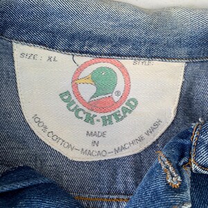 May include: Close-up of a blue denim jacket with a white tag. The tag features the "Duck Head" logo, a green duck head inside a red circle. The tag also states "100% Cotton - Macao - Machine Wash". The jacket is size XL.