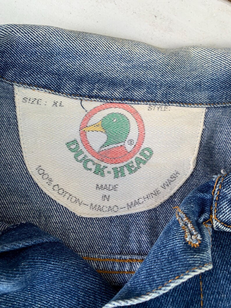 May include: Close-up of a blue denim jacket with a white tag. The tag features the "Duck Head" logo, a green duck head inside a red circle. The tag also states "100% Cotton - Macao - Machine Wash". The jacket is size XL.