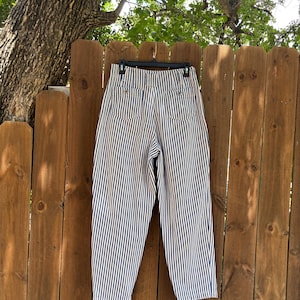 Puede incluir: Pantalones de rayas verticales azul marino y blanco, de cintura alta, colgados de una percha negra frente a una valla de madera. Los pantalones tienen dos bolsillos traseros y un corte cl&aacute;sico y relajado. Las rayas est&aacute;n espaciadas uniformemente.