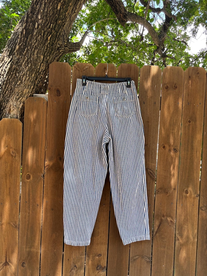 Puede incluir: Pantalones de rayas verticales azul marino y blanco, de cintura alta, colgados de una percha negra frente a una valla de madera. Los pantalones tienen dos bolsillos traseros y un corte cl&aacute;sico y relajado. Las rayas est&aacute;n espaciadas uniformemente.