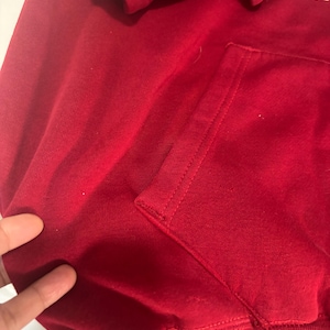 May include: Close-up of a red hooded sweatshirt with a kangaroo pocket. The hood is visible at the top, and the pocket is on the front. The fabric appears to be a soft, knit material.