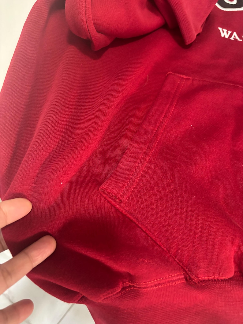 May include: Close-up of a red hooded sweatshirt with a kangaroo pocket. The hood is visible at the top, and the pocket is on the front. The fabric appears to be a soft, knit material.