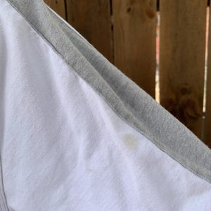 Puede incluir: Camiseta de estilo ragl&aacute;n blanca y gris. La camiseta tiene un cuerpo blanco con mangas grises y un cuello gris. Las costuras son visibles. Hay una mancha en la tela blanca.