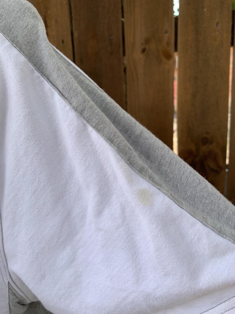 Puede incluir: Camiseta de estilo ragl&aacute;n blanca y gris. La camiseta tiene un cuerpo blanco con mangas grises y un cuello gris. Las costuras son visibles. Hay una mancha en la tela blanca.