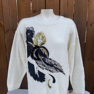 Può includere: Un maglione di lana bianco con un grande disegno floreale nero, oro e marrone. Il disegno presenta un motivo a pizzo e paillettes.