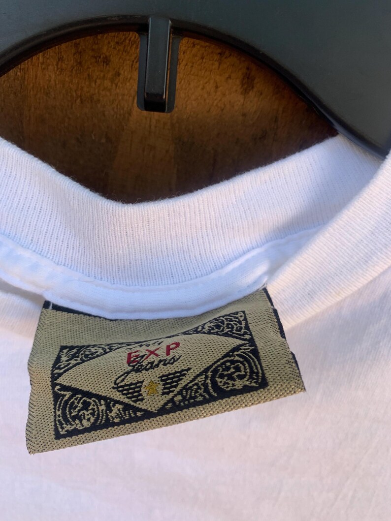 Pu&ograve; includere: Primo piano di un colletto di una maglietta bianca con un'etichetta. L'etichetta &egrave; beige con disegni neri ornati e la scritta "EXP Jeans". La maglietta &egrave; appesa a una gruccia nera su uno sfondo marrone.