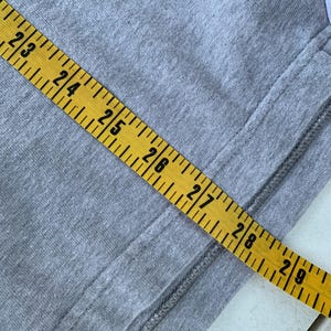 Puede incluir: Primer plano de una tela gris con una cinta m&eacute;trica amarilla. La cinta muestra medidas de 58,4 cm a 73,7 cm. Una costura recorre el lado derecho de la tela.