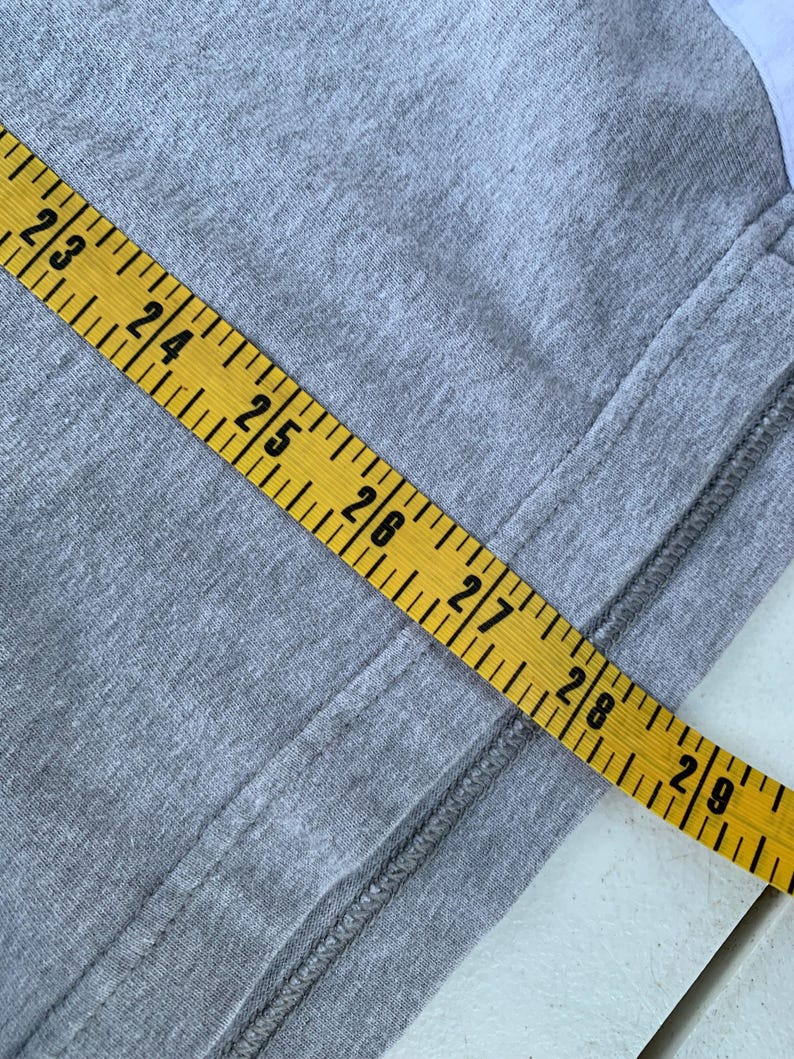 Puede incluir: Primer plano de una tela gris con una cinta m&eacute;trica amarilla. La cinta muestra medidas de 58,4 cm a 73,7 cm. Una costura recorre el lado derecho de la tela.