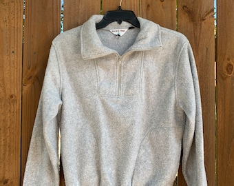 May include: A gray fleece pullover with a zipper and a Pacific Trail logo. The sweater is hanging on a black hanger against a wooden fence.