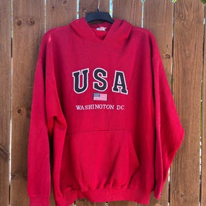 May include: A red hooded sweatshirt with the word "USA" in white block letters and a small American flag graphic. Below the text is "WASHINGTON DC" in white. The sweatshirt has a front pocket and is hanging on a wooden fence.