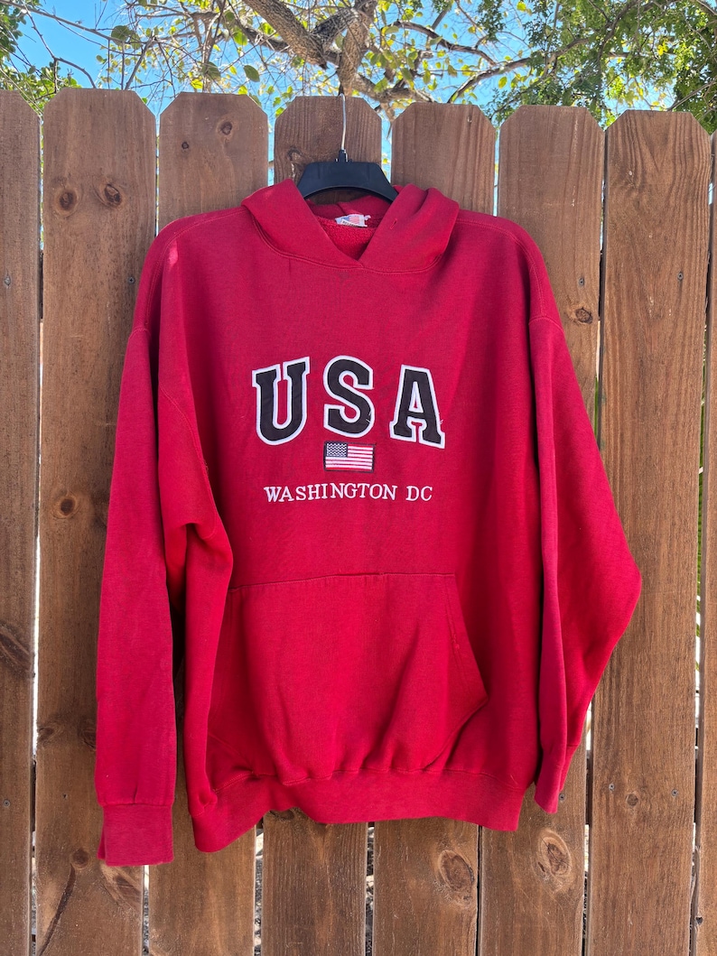 May include: A red hooded sweatshirt with the word "USA" in white block letters and a small American flag graphic. Below the text is "WASHINGTON DC" in white. The sweatshirt has a front pocket and is hanging on a wooden fence.
