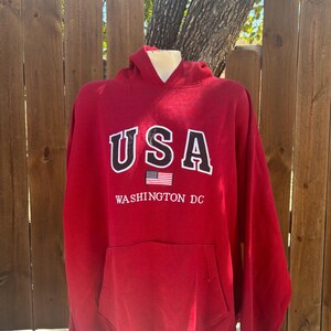 May include: A red hooded sweatshirt with the word "USA" in white block letters outlined in black. Below the text is a small American flag and the words "WASHINGTON DC" in white. The sweatshirt has a front pocket.