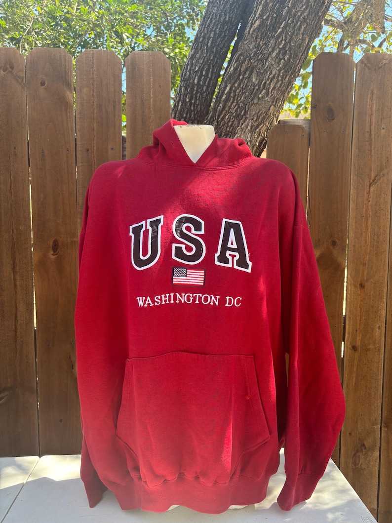 May include: A red hooded sweatshirt with the word "USA" in white block letters outlined in black. Below the text is a small American flag and the words "WASHINGTON DC" in white. The sweatshirt has a front pocket.