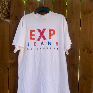 Pu&ograve; includere: T-shirt bianco con la scritta "EXP JEANS BY EXPRESS" in un gradiente di rosso, arancione, blu e viola. La maglietta &egrave; appesa a una gruccia nera contro una staccionata di legno.
