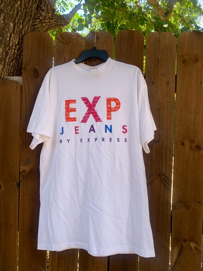 Pu&ograve; includere: T-shirt bianco con la scritta "EXP JEANS BY EXPRESS" in un gradiente di rosso, arancione, blu e viola. La maglietta &egrave; appesa a una gruccia nera contro una staccionata di legno.