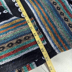 Pode incluir: T&ecirc;xtil tecido colorido com um padr&atilde;o listrado em preto, branco, vermelho, azul e amarelo. Uma fita m&eacute;trica amarela &eacute; colocada sobre o tecido, mostrando medidas em cent&iacute;metros. O t&ecirc;xtil tem uma apar&ecirc;ncia texturizada.