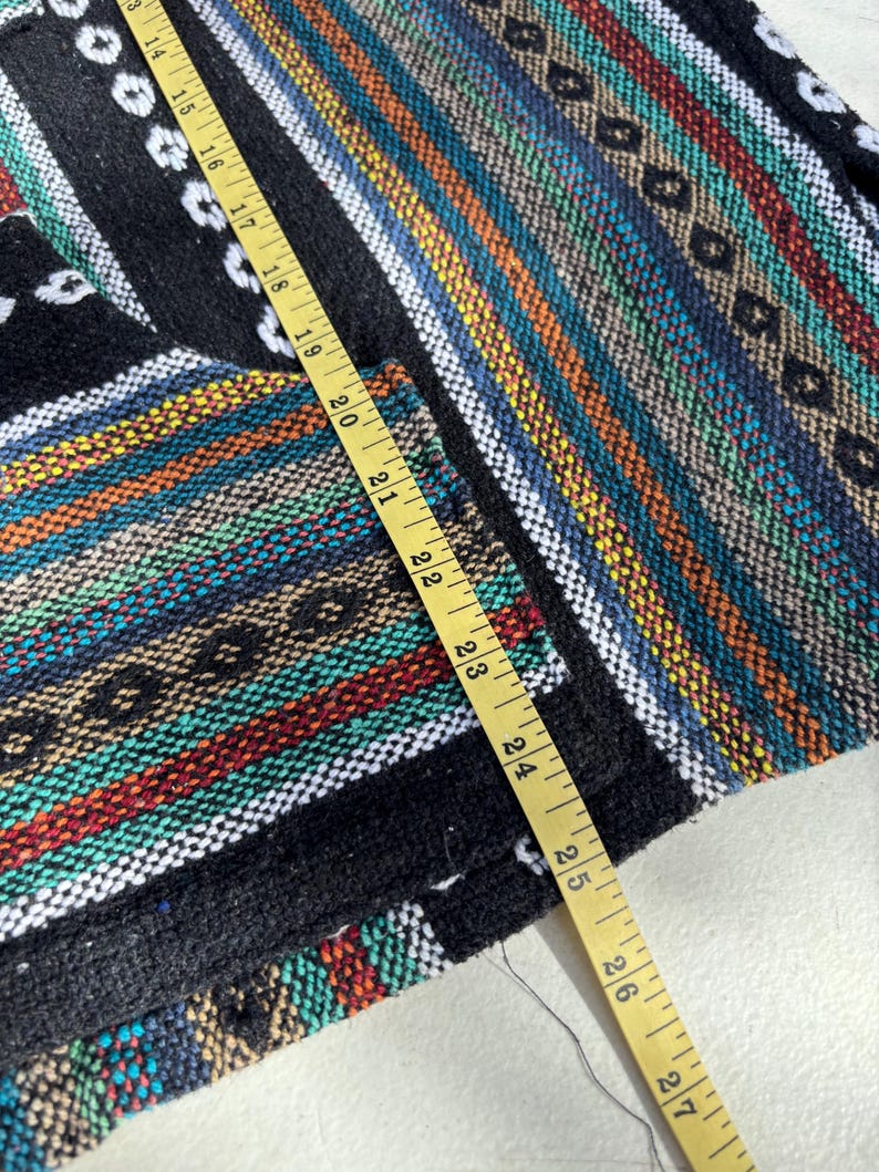 Pode incluir: T&ecirc;xtil tecido colorido com um padr&atilde;o listrado em preto, branco, vermelho, azul e amarelo. Uma fita m&eacute;trica amarela &eacute; colocada sobre o tecido, mostrando medidas em cent&iacute;metros. O t&ecirc;xtil tem uma apar&ecirc;ncia texturizada.