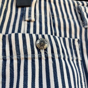 Puede incluir: Primer plano de unos pantalones vaqueros a rayas azules y blancas. Los pantalones tienen un bot&oacute;n plateado con el logotipo de Calvin Klein en el bolsillo. Las rayas son verticales y est&aacute;n espaciadas uniformemente. Los pantalones est&aacute;n colgados de una percha negra.