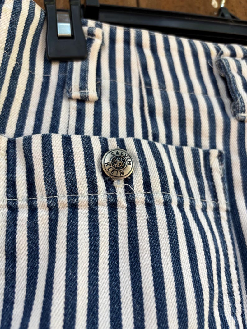 Puede incluir: Primer plano de unos pantalones vaqueros a rayas azules y blancas. Los pantalones tienen un bot&oacute;n plateado con el logotipo de Calvin Klein en el bolsillo. Las rayas son verticales y est&aacute;n espaciadas uniformemente. Los pantalones est&aacute;n colgados de una percha negra.