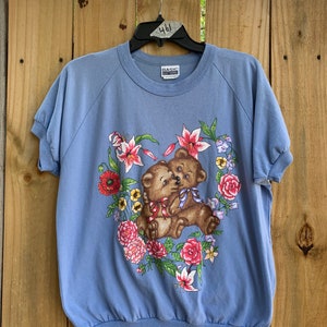 May include: A pale blue short-sleeved sweatshirt with a floral heart design. The heart features two brown teddy bears and various flowers in pink, red, and yellow. The sweatshirt is hanging on a black hanger.
