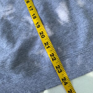 May include: Close-up of a gray sweatshirt with a yellow measuring tape laid across it. The tape shows measurements from 14 to 26 inches. The sweatshirt has a ribbed hem.