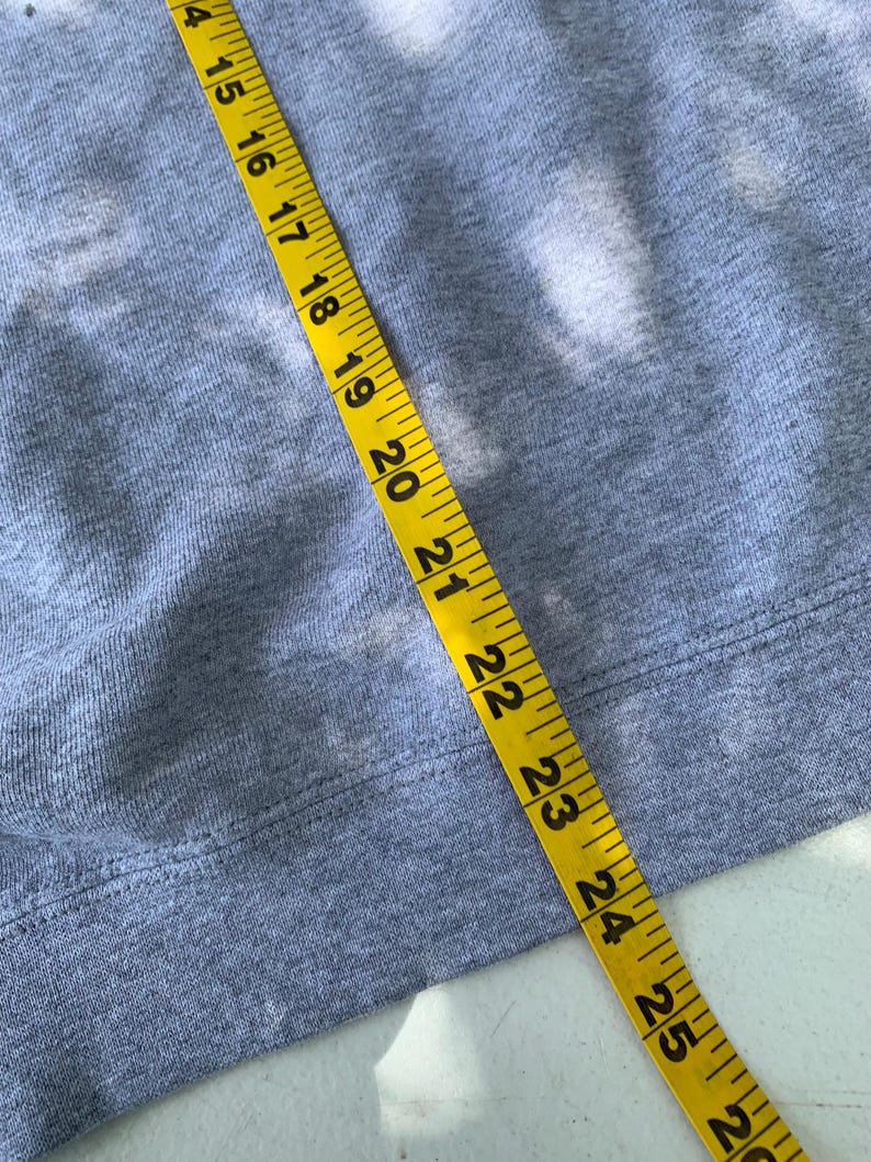 May include: Close-up of a gray sweatshirt with a yellow measuring tape laid across it. The tape shows measurements from 14 to 26 inches. The sweatshirt has a ribbed hem.