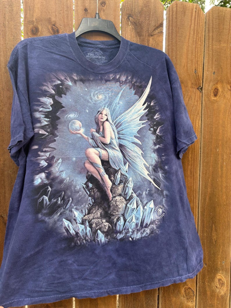 May include: Navy blue t-shirt featuring a fantasy art print of a fairy with blue wings, holding a glowing orb. The fairy is seated on a rock formation with blue crystals. The shirt is hanging on a black hanger.