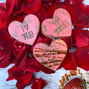 May include: Three pink heart-shaped soaps with glitter and messages "I Love You", "Too Sweet", and "Cutie Pie" on a white marble surface with red rose petals. A red strawberry with caramel sauce is in the lower right corner.