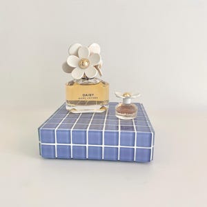 May include: Two perfume bottles, one large and one small, sit on a blue tiled surface. The larger bottle is labeled "DAISY" and has a white daisy-shaped cap. The smaller bottle, labeled "DAISY LOVE", also has a daisy cap. The background is plain white.