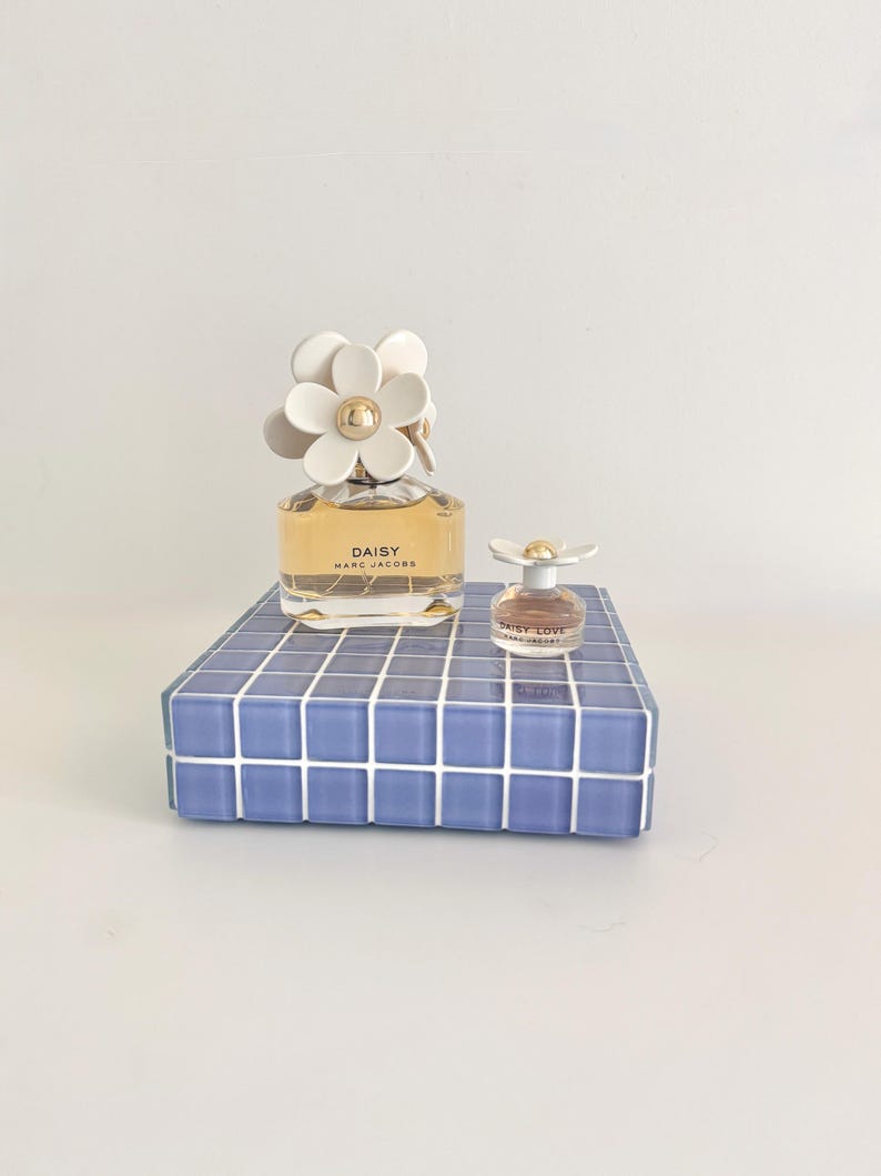 May include: Two perfume bottles, one large and one small, sit on a blue tiled surface. The larger bottle is labeled "DAISY" and has a white daisy-shaped cap. The smaller bottle, labeled "DAISY LOVE", also has a daisy cap. The background is plain white.