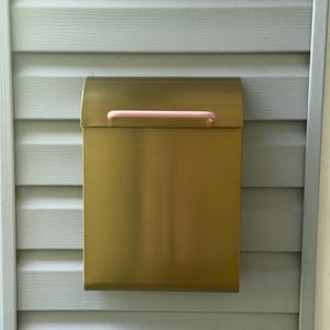 May include: A rectangular wall-mounted mailbox with a metallic gold finish and a smooth, matte texture. The mailbox features a curved top lid and a horizontal, cylindrical handle in a soft, pale pink color centered on the front flap.