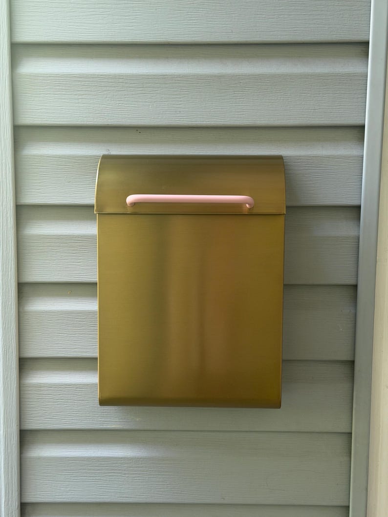 May include: A rectangular wall-mounted mailbox with a metallic gold finish and a smooth, matte texture. The mailbox features a curved top lid and a horizontal, cylindrical handle in a soft, pale pink color centered on the front flap.