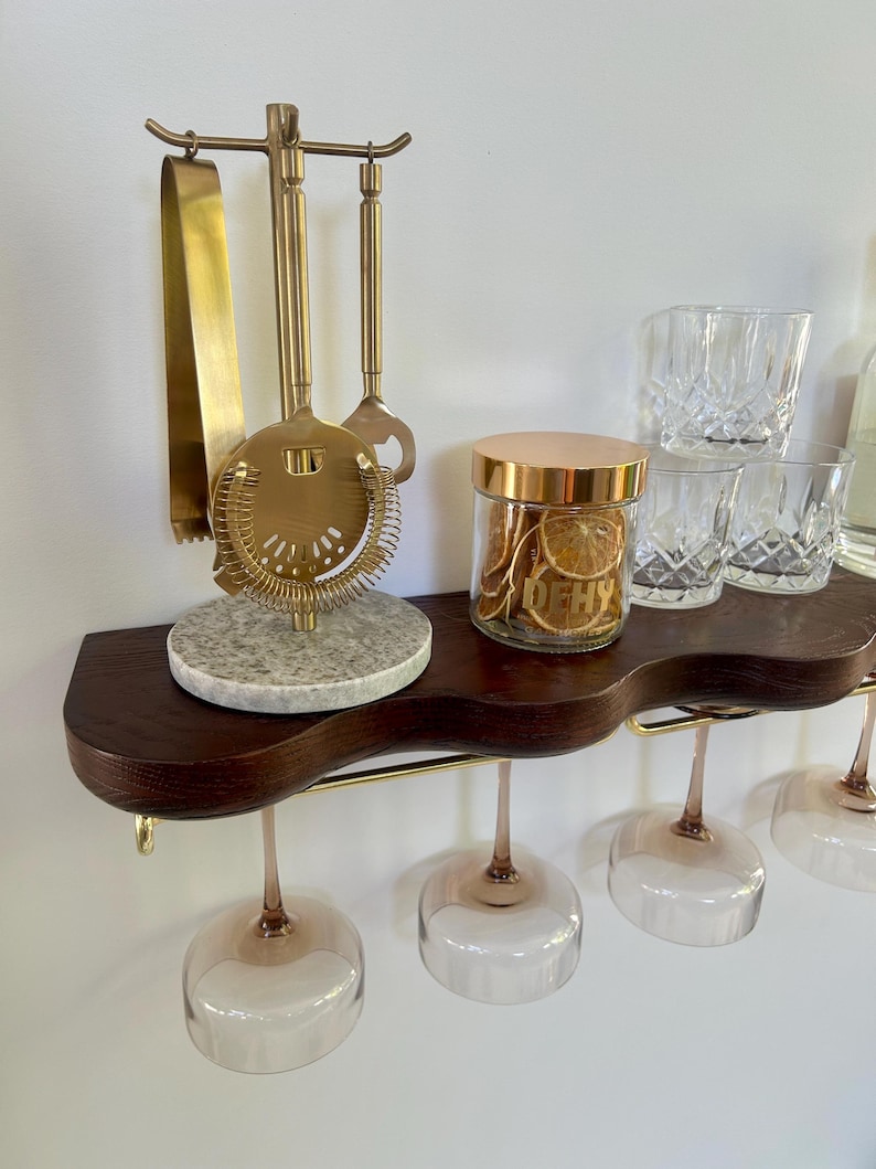 May include: A dark wood wall shelf with a wavy front edge and a gold metal rail holding four stemmed glasses. On the shelf sits a gold bar tool set on a speckled gray stone base, a glass jar with a gold lid containing dried citrus slices labeled DEHY.