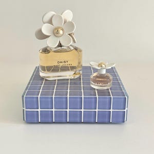 May include: Two bottles of Daisy Marc Jacobs perfume. The larger bottle has a white daisy flower cap and sits on a blue tiled platform. A smaller bottle of Daisy Love perfume is also present.