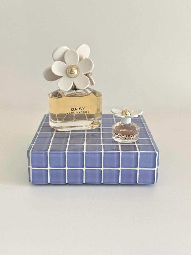 May include: Two bottles of Daisy Marc Jacobs perfume. The larger bottle has a white daisy flower cap and sits on a blue tiled platform. A smaller bottle of Daisy Love perfume is also present.