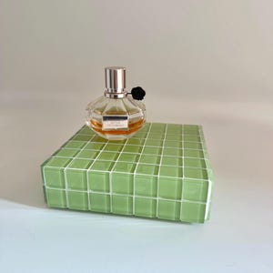 May include: A clear, round perfume bottle with a silver cap and black flower accent, labeled "Nectar Viktor & Rolf", sits atop a green tiled platform. The perfume has a light amber hue. The platform is a rectangular shape.