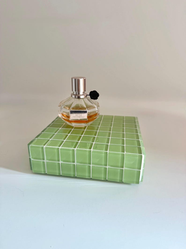 May include: A clear, round perfume bottle with a silver cap and black flower accent, labeled "Nectar Viktor & Rolf", sits atop a green tiled platform. The perfume has a light amber hue. The platform is a rectangular shape.