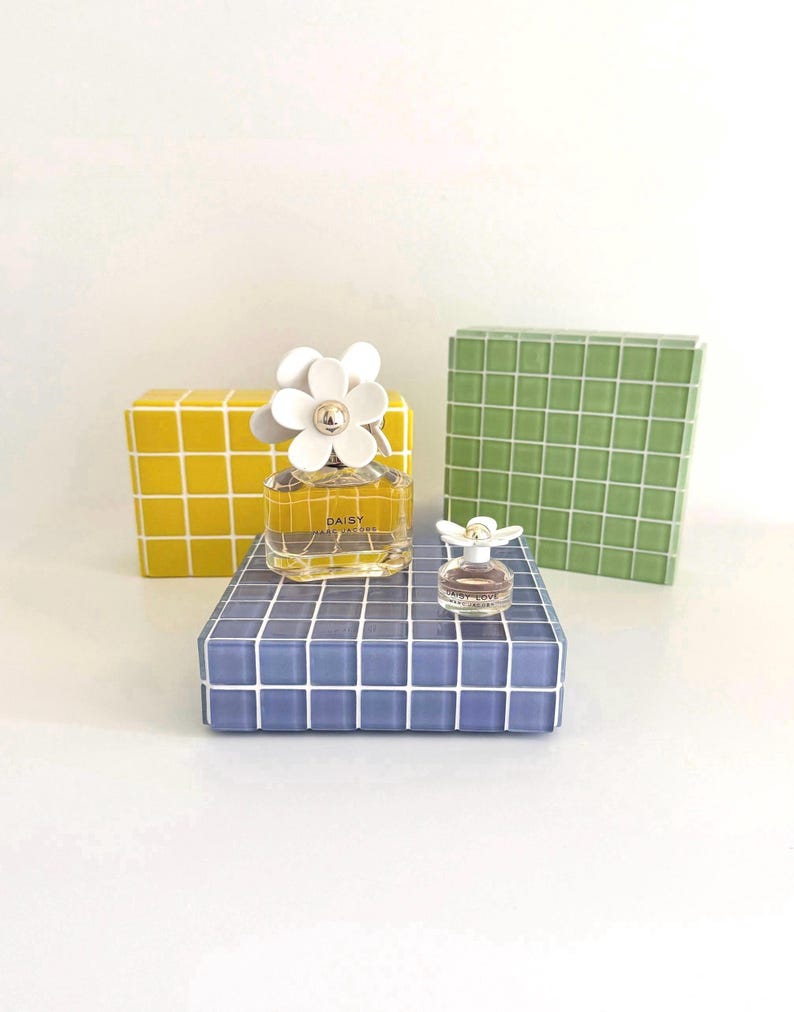 May include: Two bottles of Daisy Marc Jacobs perfume, one large and one small, with white flower-shaped caps. The bottles sit on a blue tiled surface, with yellow and green tiled blocks in the background. The perfume bottles are clear.