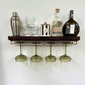 May include: A dark wood wall-mounted shelf with a gold-toned metal rack holding four green-tinted wine glasses. The shelf displays a stainless steel shaker, three crystal tumblers, a black and white striped jar, and two glass liquor bottles.