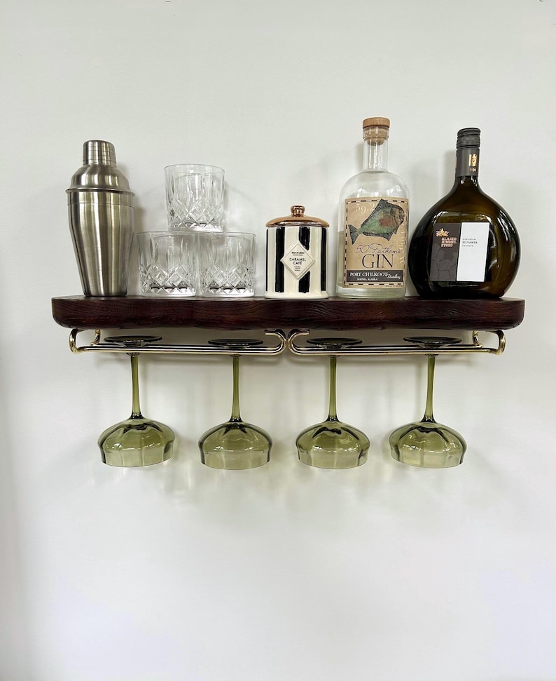 May include: A dark wood wall-mounted shelf with a gold-toned metal rack holding four green-tinted wine glasses. The shelf displays a stainless steel shaker, three crystal tumblers, a black and white striped jar, and two glass liquor bottles.