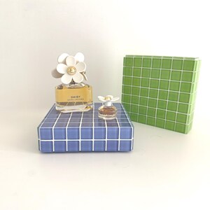 May include: A perfume bottle with a white daisy flower cap and the word "DAISY" printed on the bottle. A miniature perfume bottle with a daisy cap sits on a blue tiled surface. A green tiled box is in the background.