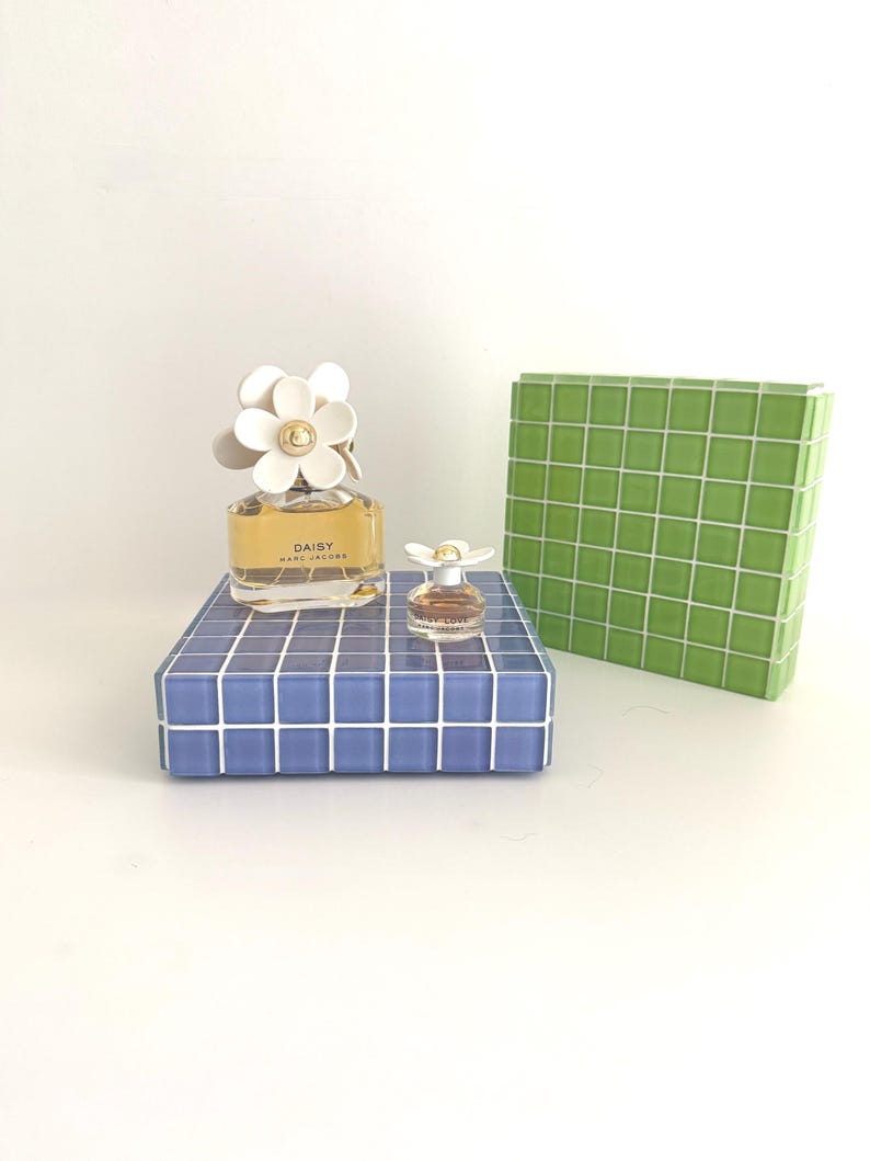 May include: A perfume bottle with a white daisy flower cap and the word "DAISY" printed on the bottle. A miniature perfume bottle with a daisy cap sits on a blue tiled surface. A green tiled box is in the background.