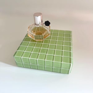 May include: A clear, faceted perfume bottle with a silver cap and black flower accent sits atop a green tiled block. The perfume has a light amber hue. The block is a light green color with white grout lines.