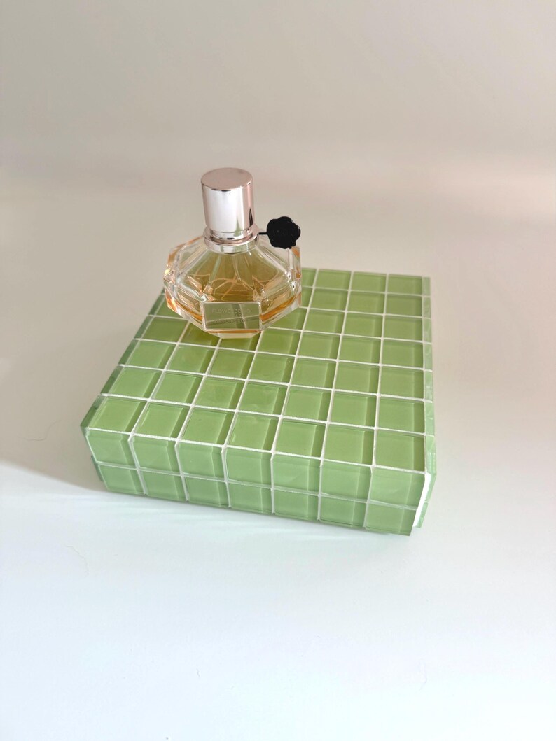 May include: A clear, faceted perfume bottle with a silver cap and black flower accent sits atop a green tiled block. The perfume has a light amber hue. The block is a light green color with white grout lines.