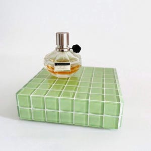 May include: A clear perfume bottle with a silver cap and black flower accent sits on a green tiled platform. The perfume bottle contains a light amber liquid. The platform is a rectangular shape with a grid pattern.