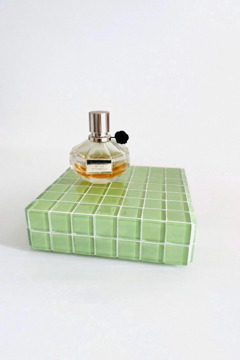 May include: A clear perfume bottle with a silver cap and black flower accent sits on a green tiled platform. The perfume bottle contains a light amber liquid. The platform is a rectangular shape with a grid pattern.