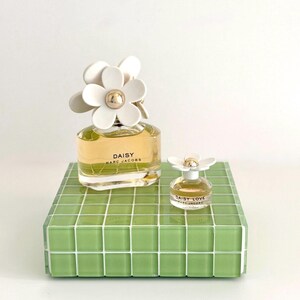 May include: Two perfume bottles, one large and one small, sit on a green tiled surface. The larger bottle is labeled "DAISY MARC JACOBS" and has a white flower-shaped cap. The smaller bottle, "DAISY LOVE MARC JACOBS", also has a flower cap.