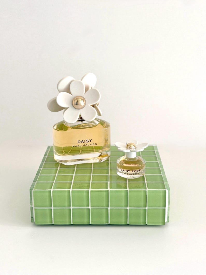 May include: Two perfume bottles, one large and one small, sit on a green tiled surface. The larger bottle is labeled "DAISY MARC JACOBS" and has a white flower-shaped cap. The smaller bottle, "DAISY LOVE MARC JACOBS", also has a flower cap.