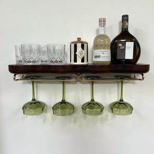 May include: A dark wood wall-mounted shelf with a gold metal rack holding four green-tinted stemmed glasses. The shelf top displays three clear crystal tumblers, a black and white striped canister with a gold lid, and two glass bottles.